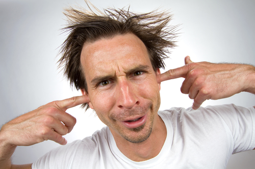 Scruffy unpleasant looking man with a silly facial expression and unruly hair puts his fingers in his ears so that he can not hear.